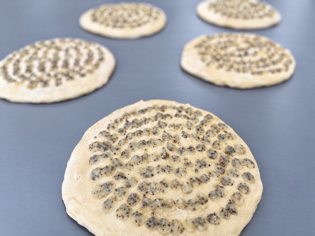 Flatbread bases with separate dots of herb sauce deposited in rings, on a conveyor belt