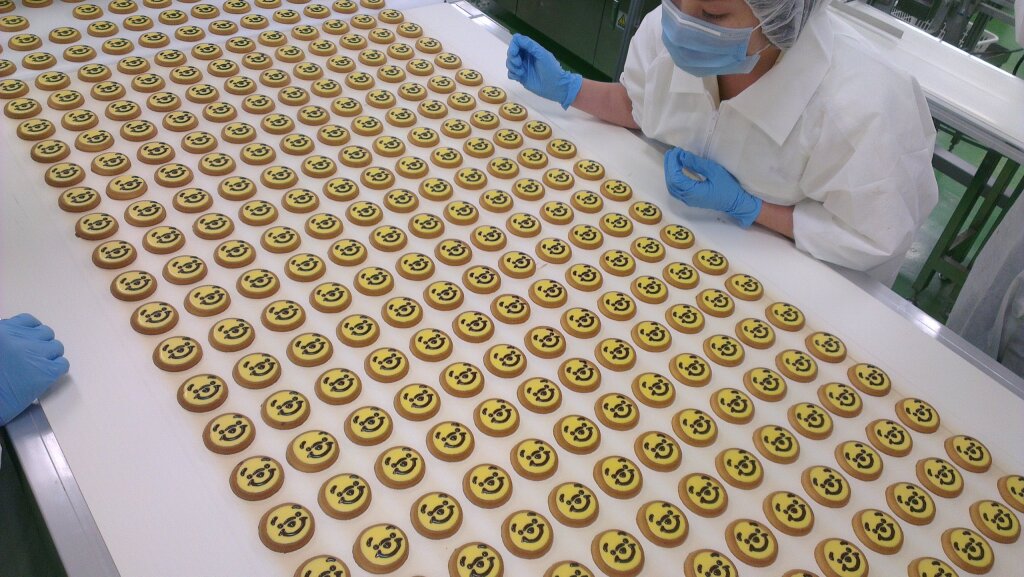 Rows of biscuits with smiley decorations on a conveyor, checked by an operator