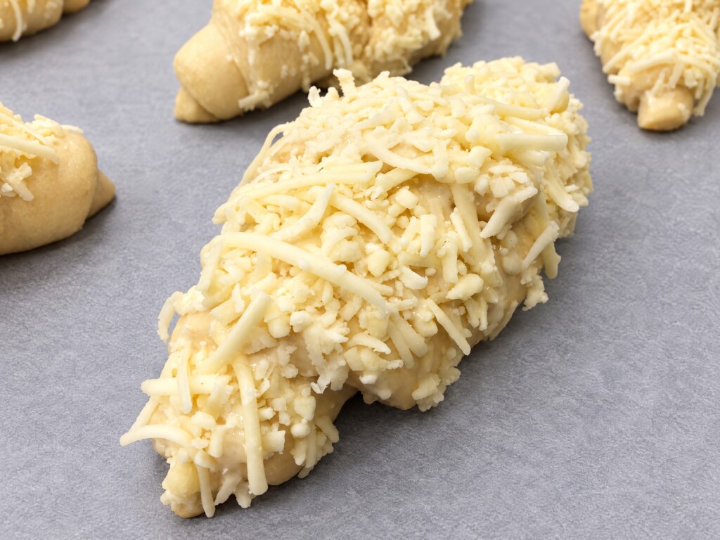 Unbaked croissant covered with grated cheese on a glossy sauce layer